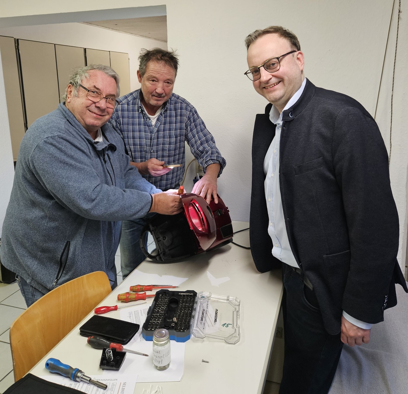MdL Meyer (r.) sieht den Handwerkern (v. l.) Herbert und Peter beim Reparieren eines Staubsaugers zu.